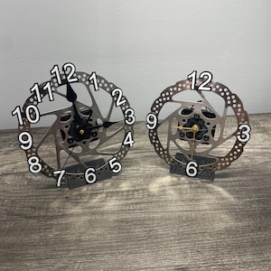 May include: Two silver bicycle disc brake clocks with black hands and white numbers. The clocks are on a wood surface.