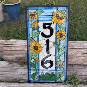 May include: A stained glass house number sign with the number 516 in black on a white background. The sign is surrounded by a mosaic of sunflowers and leaves in shades of yellow, green, and blue.