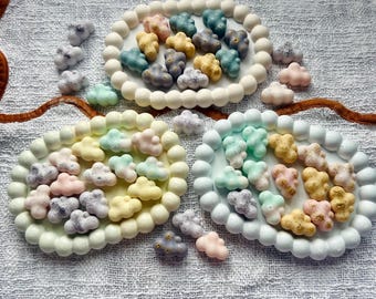 Könnte beinhalten: Drei ovale Tabletts mit wolkenförmigen Süßigkeiten in Pastellfarben. Jedes Tablett hat einen weißen Perlenrand. Die Süßigkeiten sind in Rosa, Gelb, Blau und Grau gehalten, einige mit goldenen Akzenten. Auf einer weißen, strukturierten Oberfläche.