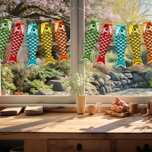 May include: A string of fifteen decorative carp-shaped windsocks, known as koinobori, hanging in front of a window. The fish are arranged in three repeating sets of five, featuring green, red, blue, yellow, and orange colors with scale patterns.