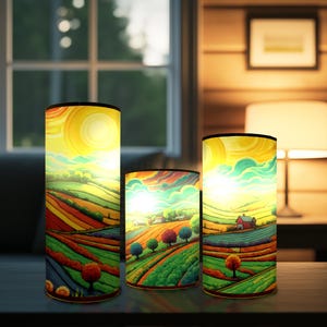 May include: Three cylindrical lamps featuring a vibrant landscape illustration with rolling hills, green fields, and a bright sun. The artwork displays a colourful rural scene with trees and a small house, illuminated from within to highlight the yellow, orange, and green tones.