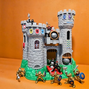 May include: A grey plastic toy castle with two turrets, flags, and a drawbridge. Several toy knights in gold and black armour are positioned around the castle. The castle sits on a green base.