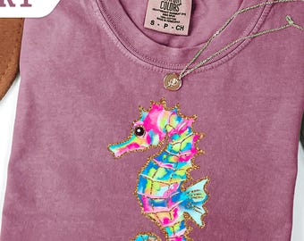 May include: Berry-colored t-shirt with a colorful seahorse design. The seahorse is in shades of pink, blue, green, and yellow, with gold glitter accents. A gold necklace with a pendant rests on the shirt. The word "BERRY" is in a white banner.