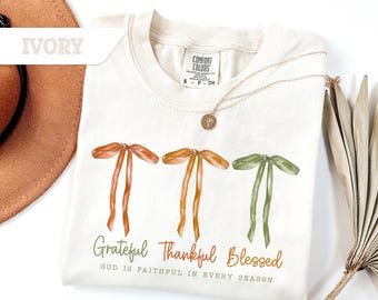 May include: Ivory-colored t-shirt with three watercolor bows in shades of orange and green. The words "Grateful, Thankful, Blessed" are printed below the bows, with the phrase "GOD IS FAITHFUL IN EVERY SEASON" underneath.