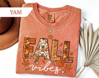 May include: A coral-colored t-shirt with the words "FALL vibes" in a decorative font. The word "FALL" is filled with fall-themed illustrations of leaves, acorns, and pumpkins. A gold necklace and a brown hat are also visible.