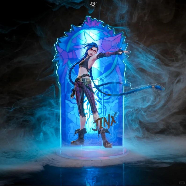 May include: A blue and purple stained glass-style acrylic stand with a figure of a female character with blue hair and a gun. The character is standing on a clear acrylic base. The stand has the word "JINX" written on it.