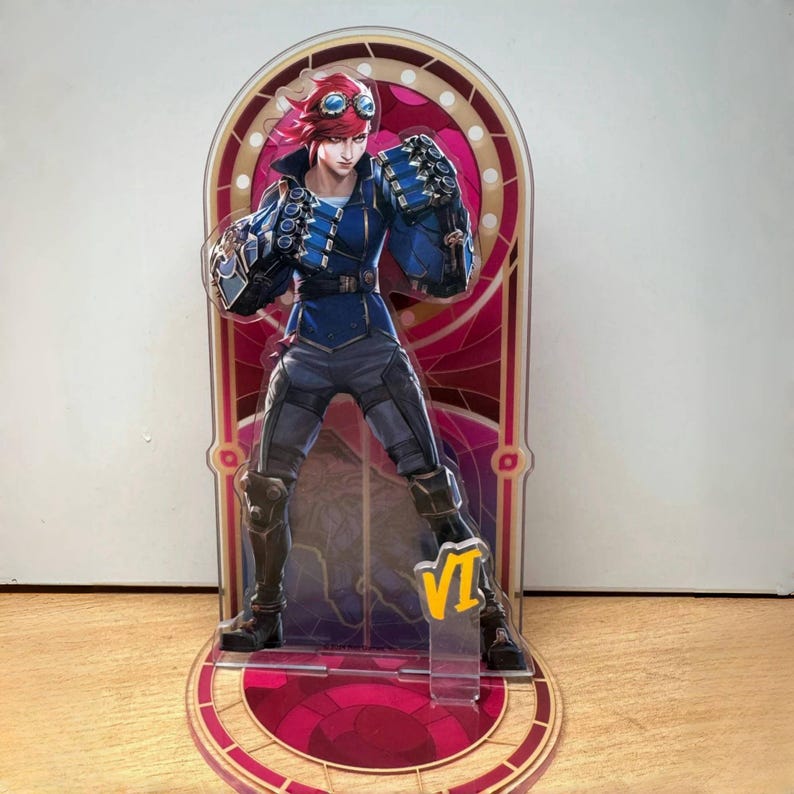 May include: A red-haired character with goggles and a blue jacket stands on a clear acrylic stand with a red and gold design. The character is holding a weapon in their right hand. The stand has the number "VI" on it.