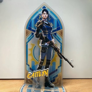 May include: A blue and gold character stand featuring a female figure with blue hair holding a rifle. The character's name, "Caitlyn", is printed in yellow on the stand.