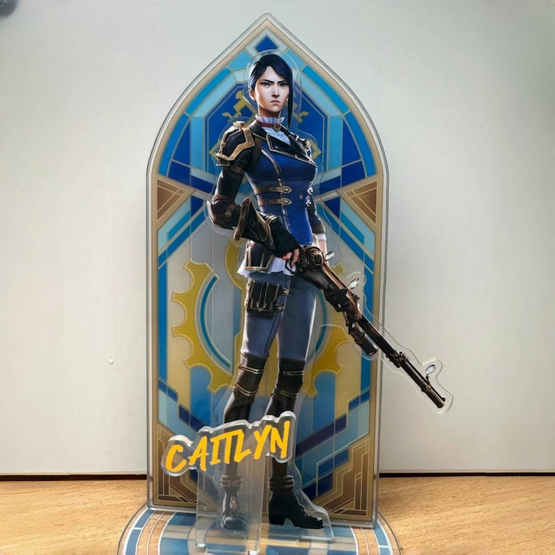 May include: A blue and gold character stand featuring a female figure with blue hair holding a rifle. The character's name, "Caitlyn", is printed in yellow on the stand.