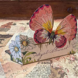 May include: A decorative paper butterfly with pink, yellow, and purple wings, perched atop a box adorned with daisies and the name "Hazel Stewart." The box is set on a vintage paper background.
