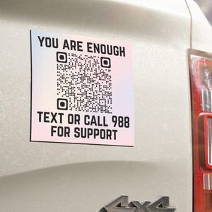 以下が含まれることがあります： 黒い文字でYOU ARE ENOUGH、中央に白黒のQRコード、黒い文字でTEXT OR CALL 988 FOR SUPPORTと書かれた正方形の白いマグネット。マグネットは4x4のエンブレムが見える白い車両のテールゲートに取り付けられています。