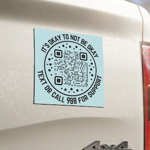 May include: A square light blue sticker applied to a white vehicle tailgate. The sticker features a central QR code surrounded by black text that reads: IT'S OKAY TO NOT BE OKAY, TEXT OR CALL 988 FOR SUPPORT, with small decorative dots.