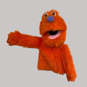 May include: An orange puppet with a fuzzy texture, open mouth, and a pink interior. It has a blue nose and two large, round eyes. The puppet's arm is extended to the left, suggesting a gesture.