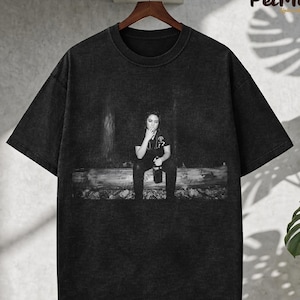 Könnte beinhalten: Eine weiße Keramik-Tasse mit schwarzem Henkel und dem Text "But First, Coffee" in fetter, schwarzer Schrift. Die Tasse steht vor einem verschwommenen Hintergrund, der eine gemütliche Umgebung andeutet.