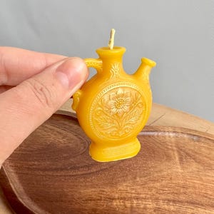 May include: A yellow beeswax candle shaped like a small, flat-sided vessel with a central circular relief featuring a sunflower design. The candle has a single wick at the top, a small handle on the side, and a secondary spout-like opening on the right.