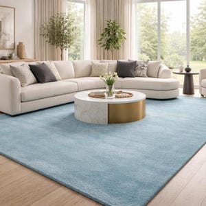 May include: A modern living room featuring a large, light blue rug, a sectional sofa, and a round coffee table. The sofa is cream-colored, and the coffee table has a marble top and gold base. The room has large windows and hardwood floors.