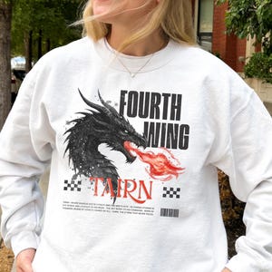 K&ouml;nnte beinhalten: Wei&szlig;es Sweatshirt mit einem schwarzen Drachenmotiv, der rote Flammen speit, mit dem Text "FOURTH WING" und "TAIRN". Das Design beinhaltet ein Schachbrettmuster und Text am unteren Rand. Es ist ein lang&auml;rmeliges Rundhals-Sweatshirt.