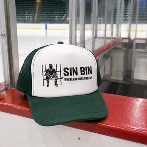 Könnte beinhalten: Eine grüne und weiße Trucker-Mütze mit dem Text "SIN BIN WHERE BAD BOYS COOL OFF" und einer schwarzen Grafik eines Hockeyspielers. Die Mütze liegt auf einer roten Oberfläche.