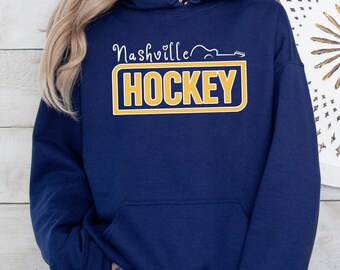 May include: Navy blue hoodie with a front graphic. The graphic reads "Nashville HOCKEY" in white and yellow lettering. The word "Nashville" is in a script font, and the word "HOCKEY" is in a bold, block font. The hoodie has a front pocket and a drawstring hood.