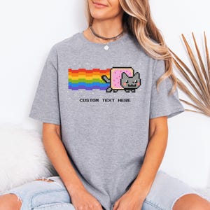 May include: A gray t-shirt featuring a pixelated Nyan Cat design with a rainbow trail. The cat has a pink body and a gray face. The text "CUSTOM TEXT HERE" is printed below the graphic.