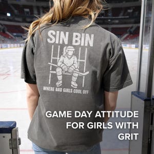 May include: Gray t-shirt with the text "SIN BIN" and a graphic of a hockey player sitting in a penalty box. The text "WHERE BAD GIRLS COOL OFF" is below the graphic. The bottom of the shirt reads "GAME DAY ATTITUDE FOR GIRLS WITH GRIT."