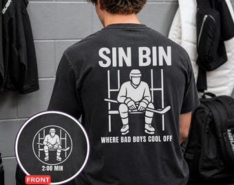 Puede incluir: Camiseta negra con el texto "SIN BIN" y un gráfico de un jugador de hockey sentado en el banquillo de penalización. La frase "WHERE BAD BOYS COOL OFF" está impresa debajo del gráfico. Un gráfico circular en la parte inferior izquierda muestra el diseño frontal.