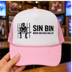 Könnte beinhalten: Rosa-weiße Trucker-Mütze mit dem Text "SIN BIN WHERE BAD GIRLS COOL OFF". Die Mütze zeigt eine schwarze Grafik eines Hockeyspielers, der auf einer Bank sitzt. Die Mütze hat einen rosa Schirm und ein Mesh-Rückenteil.