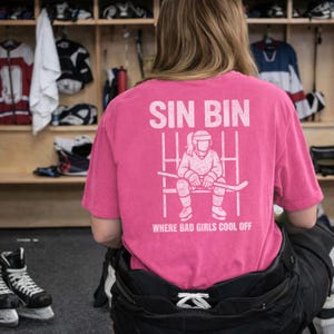 May include: A pink t-shirt with the words "SIN BIN" above a graphic of a hockey player sitting on a bench, holding a hockey stick. The text "WHERE BAD GIRLS COOL OFF" is below the graphic. The shirt is in a locker room setting.
