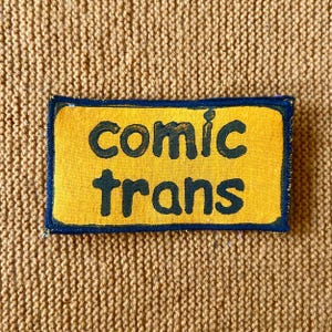 May include: A yellow fabric patch with black stitching and the words "comic trans" in black lettering.