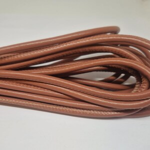 Puede incluir: Primer plano de un rollo de cordón de cuero marrón. El cordón es liso y flexible, y es perfecto para proyectos de artesanía.