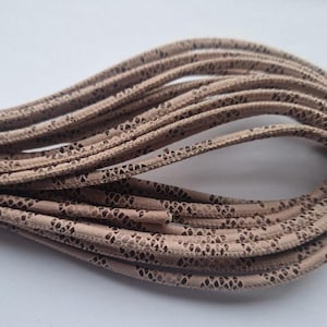 Może przedstawiać: Zbliżenie na szpulkę beżowych sznurowadeł z wzorem. Sznurowadła są wykonane z miękkiego, elastycznego materiału i mają subtelny połysk.