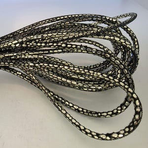 Może przedstawiać: Zbliżenie na wiązkę ozdobnych listew. Wykończenie jest czarne ze srebrnym, teksturowanym wzorem. Wykończenie jest zwinięte i wydaje się być wykonane z elastycznego materiału, odpowiedniego do rękodzieła lub zdobień.