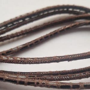 Może przedstawiać: Zbliżenie na brązowy sznurek skórzany z tkanym wzorem. Sznurek jest miękki i elastyczny, a jego powierzchnia delikatnie się błyszczy.