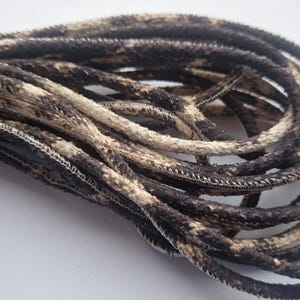 May include: Close-up of a bundle of shoelaces with a snakeskin pattern. The laces are primarily dark brown and beige, with a textured appearance. The edges are stitched with white thread.
