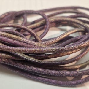 May include: Close-up of a purple and gold leather cord. The cord is braided and has a textured surface.