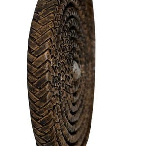 Può includere: Oggetto circolare marrone con trama intrecciata e motivo a spirale. L'oggetto ha un retro piatto e un bordo arrotondato. La trama è realizzata con strisce intrecciate, creando un motivo dettagliato. Probabilmente è un oggetto decorativo.
