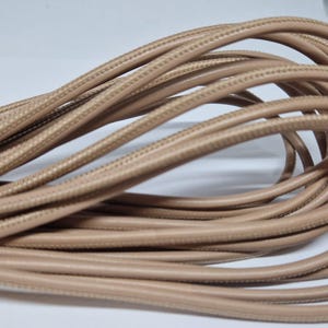 Op de afbeelding: Close-up van een bundel beige elektrische draad. De draad is gevlochten en heeft een gladde, afgeronde buitenkant. De draden zijn gelust en opgerold, waardoor een textuurpatroon ontstaat. De neutrale kleur van de draad suggereert een veelzijdig designelement.