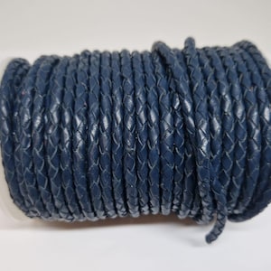 May include: A roll of navy blue braided leather cord. The cord is tightly wound and has a textured, woven appearance. The roll is white and the cord is a deep, saturated blue.