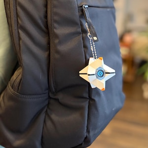 May include: A black backpack with a white, geometric, star-shaped keychain attached to the zipper. The keychain features a central blue circular emblem with a black symbol and small orange accents on the points of the star-shaped structure.