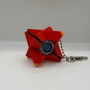 May include: A red, star-shaped geometric keychain with orange tips and a central circular eye feature. The eye is grey with a blue graphic symbol in the center. A silver-toned metal ball chain and lobster clasp are attached to the side.