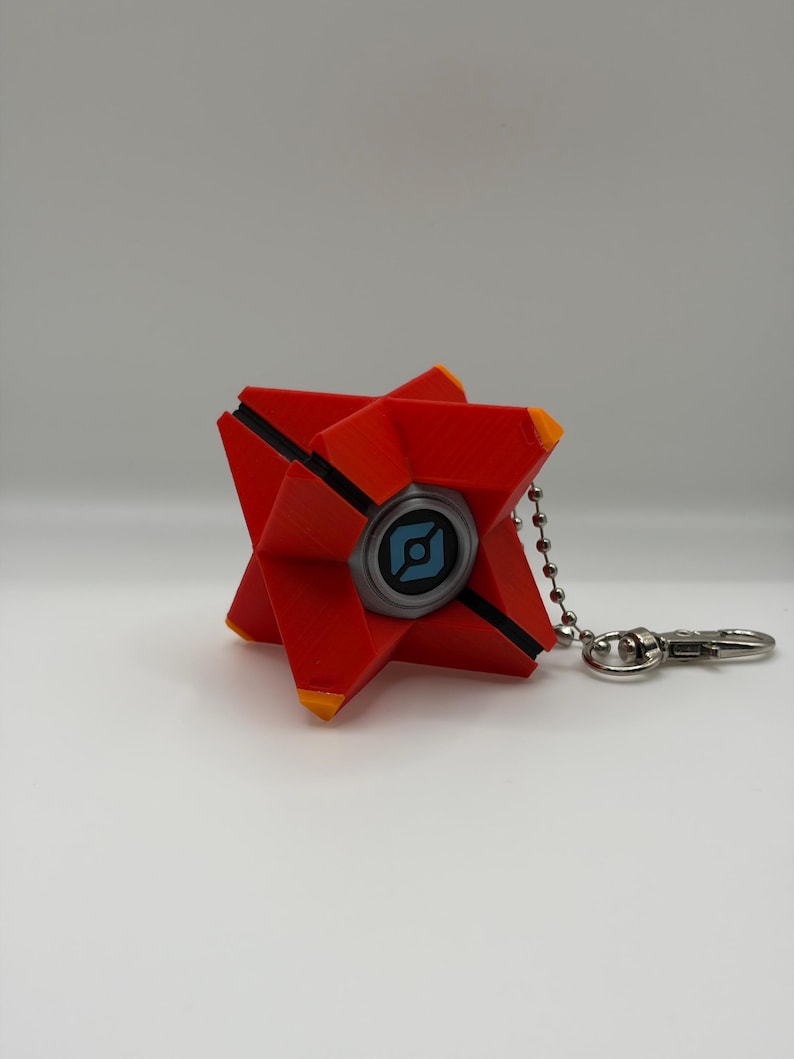 May include: A red, star-shaped geometric keychain with orange tips and a central circular eye feature. The eye is grey with a blue graphic symbol in the center. A silver-toned metal ball chain and lobster clasp are attached to the side.
