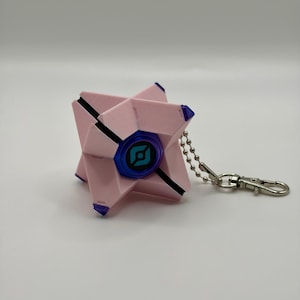 May include: A light pink, geometric, star-shaped keychain featuring a central blue circular eye with a black graphic symbol. The object has small purple accents on its points and is attached to a silver-colored metal ball chain and clasp.