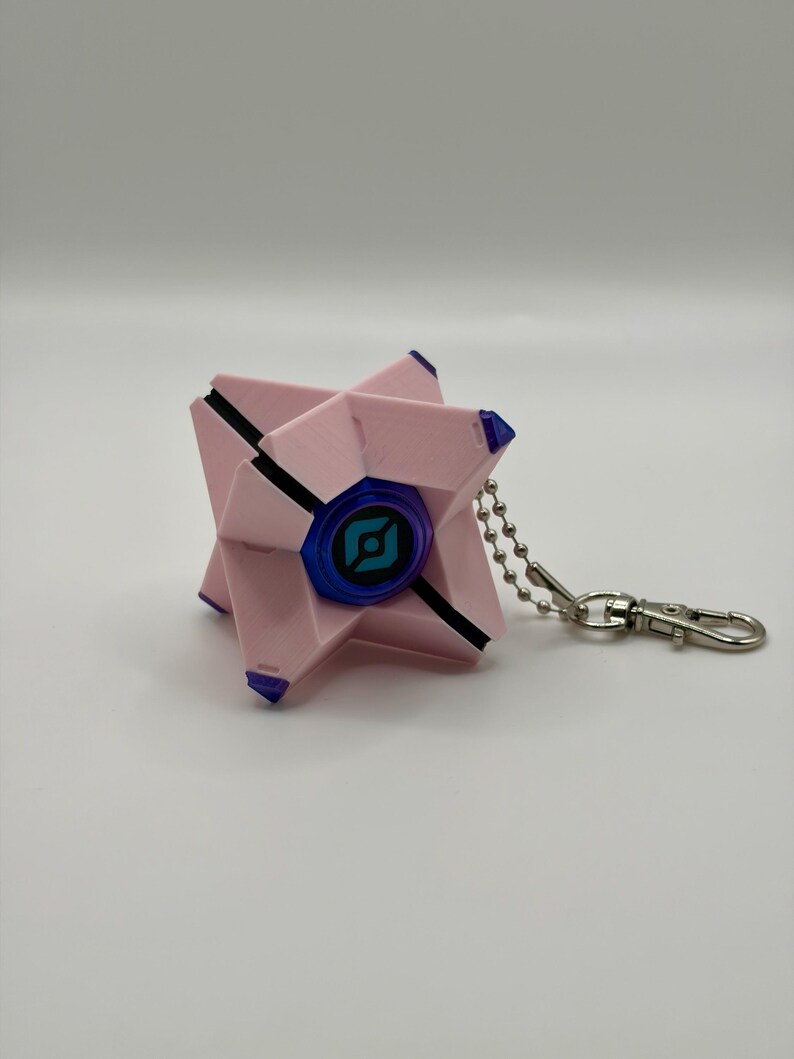 May include: A light pink, geometric, star-shaped keychain featuring a central blue circular eye with a black graphic symbol. The object has small purple accents on its points and is attached to a silver-colored metal ball chain and clasp.