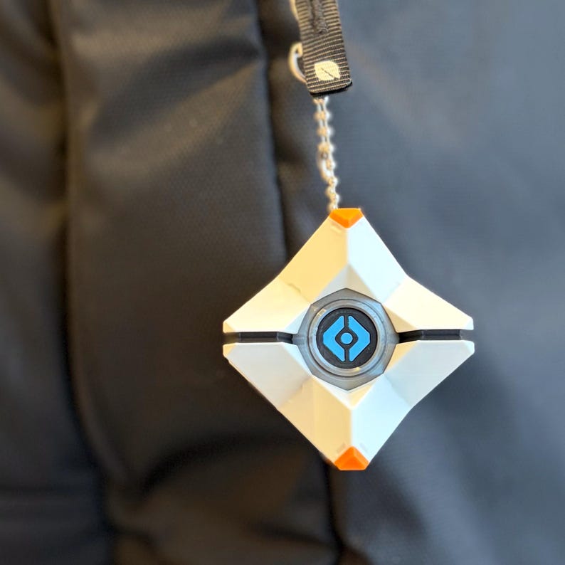 May include: A white, diamond-shaped geometric keychain featuring a central circular blue emblem with a black outline. The object has small orange accents at the top and bottom points and is attached to a silver-toned metal ball chain.