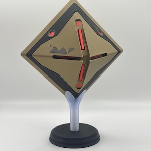 May include: A geometric, diamond-shaped object in shades of brown, black, and red, mounted on a stand. The object has a detailed design with red accents and a black border. The stand is composed of a black base, a clear stem, and a clear support.