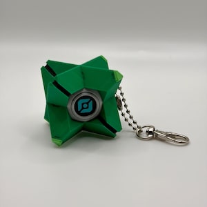 May include: A geometric green keychain featuring a central circular eye element with a blue and black graphic icon. The object has a multi-faceted, angular structure and is attached to a silver-toned metal ball chain with a lobster claw clasp.