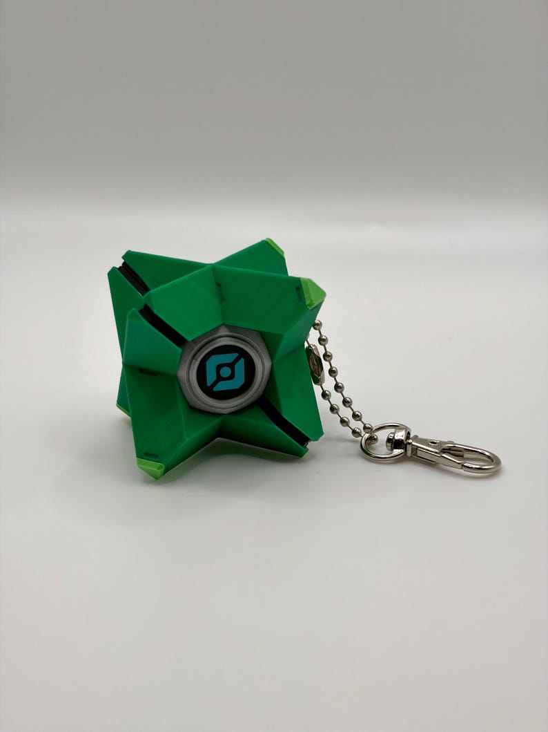 May include: A geometric green keychain featuring a central circular eye element with a blue and black graphic icon. The object has a multi-faceted, angular structure and is attached to a silver-toned metal ball chain with a lobster claw clasp.