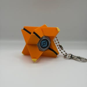 May include: A geometric orange keychain featuring a central circular element with a light blue icon on a dark background. The object has sharp, angular points with yellow tips and black recessed lines, attached to a silver metal ball chain and clasp.