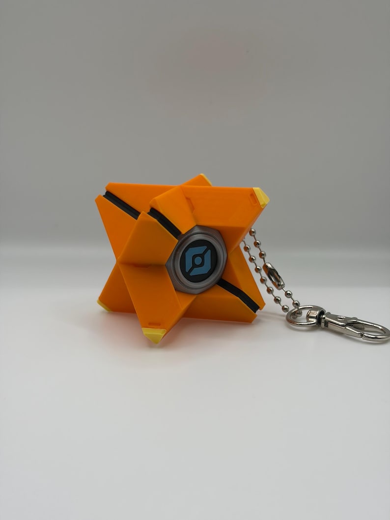 May include: A geometric orange keychain featuring a central circular element with a light blue icon on a dark background. The object has sharp, angular points with yellow tips and black recessed lines, attached to a silver metal ball chain and clasp.