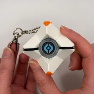 May include: A white, geometric, diamond-shaped keychain featuring orange accents at the top and bottom points. The center contains a circular gray frame surrounding a blue eye-like icon with a black center. A silver metal ball chain and clasp are attached.